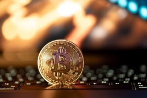 A bitcoin stands on a keyboard with an orange computer screen in the background.